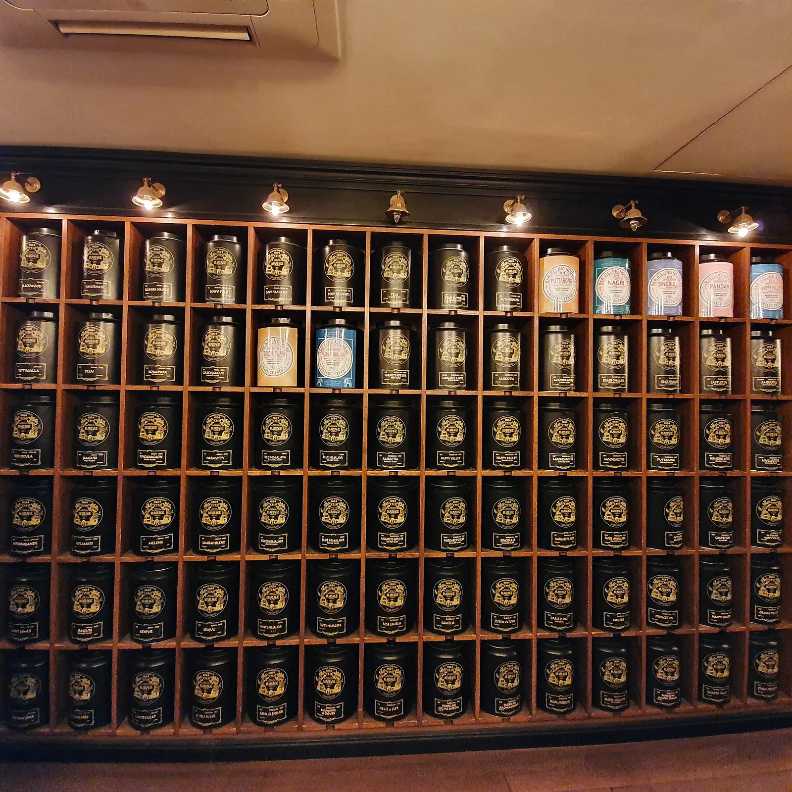 A wall of wooden cubbyholes holding black and pastel Mariage Frères tea tins at the Madeleine counter.