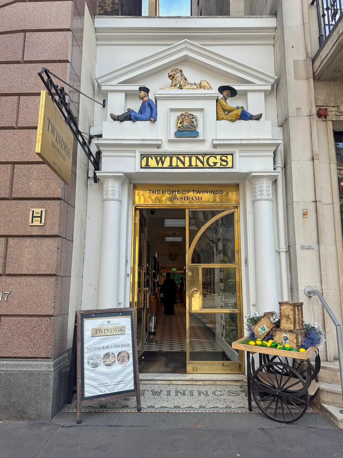 Twinings at 216 Strand in March 2026: the doorway, the TWININGS mosaic on the doorstep, a 'Tasting Bar & Tea Shop' board and a small cart of tea tins outside.